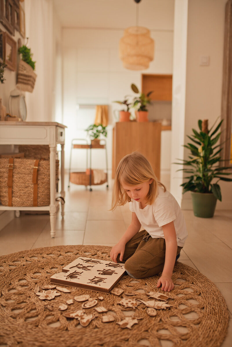 Ein kleines Kind sitzt auf einem geflochtenen Teppich und spielt mit einem Holzpuzzle, das das Leben eines Käfers darstellt. Im Hintergrund ist eine helle, gemütliche Wohnung mit Pflanzen und Möbelstücken zu sehen.