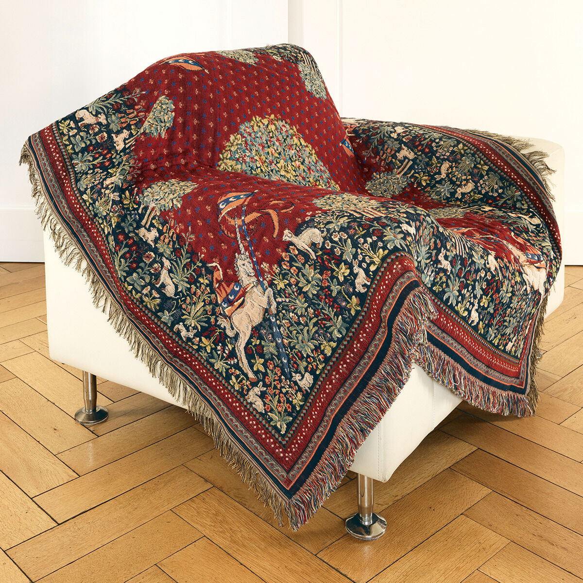 Ein kunstvoll gewebtes Gobelin-Plaid mit detailreichem Mittelaltermotiv liegt dekorativ über einem weißen Sessel. Es zeigt Einhörner in den Ecken, üppige Blumen und Tiere auf rotem und dunkelblauem Grund, umrahmt von Fransen. Elegant und ideal als Blickfang im Wohnzimmer.
