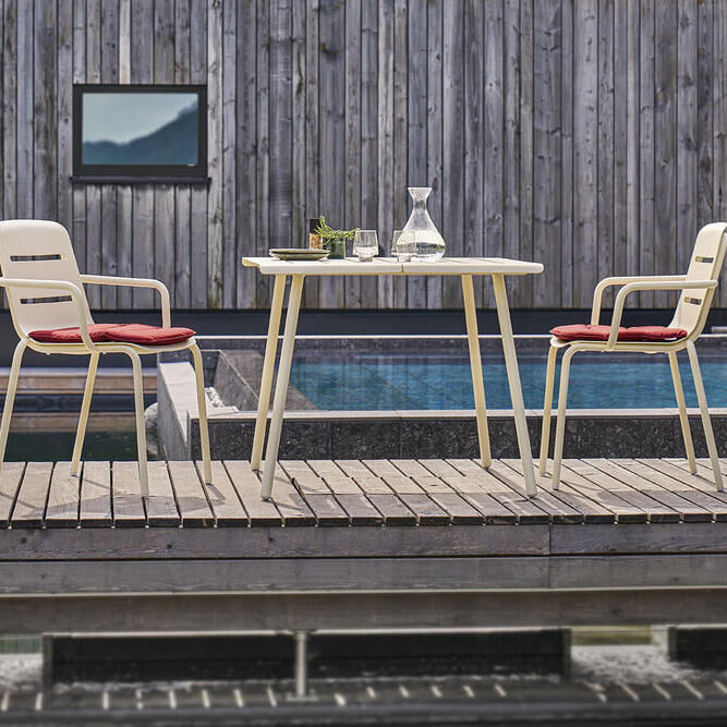 Zwei beige Stühle mit roten Kissen um einen runden weißen Tisch auf einer Terrasse, im Hintergrund ein Pool und ein Holzwand.