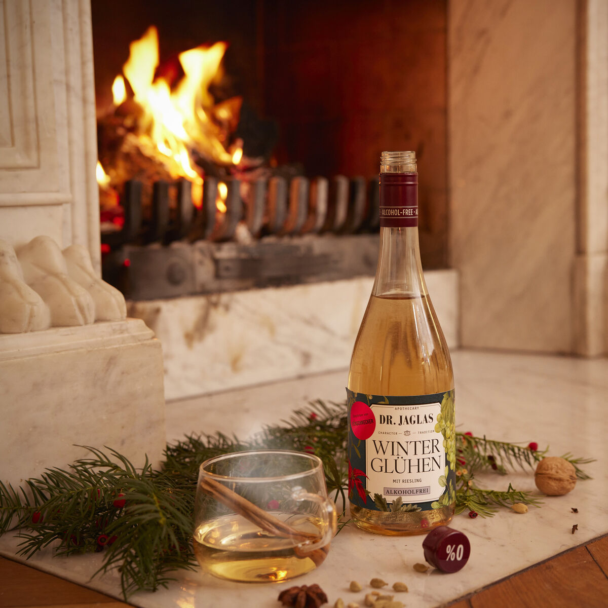Eine Flasche Winterglühen Riesling und ein Glas mit Getränk stehen auf einem Marmorkamin vor einem brennenden Kaminfeuer. Deko mit Tannenzweigen, Walnüssen und festlichen Akzenten sorgt für eine gemütliche Winter- und Weihnachtsatmosphäre.