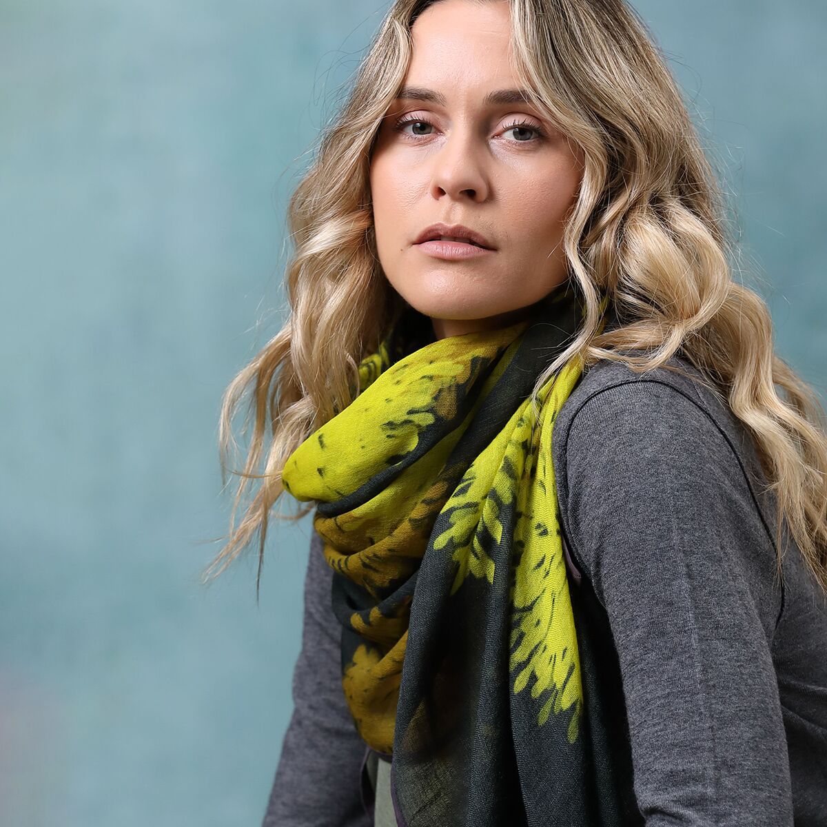 Eine Frau trägt einen gemusterten, gelben Wollschal »Carol« über einem grauen Oberteil vor blau-grünem Hintergrund. Der Schal wirkt weich, modern und eignet sich als modisches Accessoire für Damen im Herbst oder Winter.