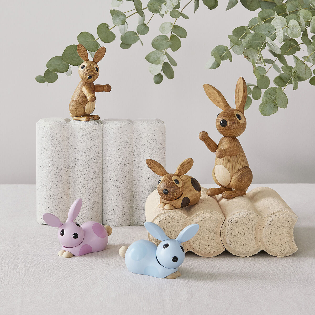 Dekorationsfiguren in Form von Hasen aus Holz, platziert auf gewellten weißen und beigen Sockeln, umgeben von grünen Eukalyptuszweigen. Zwei kleinere Hasen in Pastellrosa und Pastellblau liegen auf einer weißen Oberfläche.