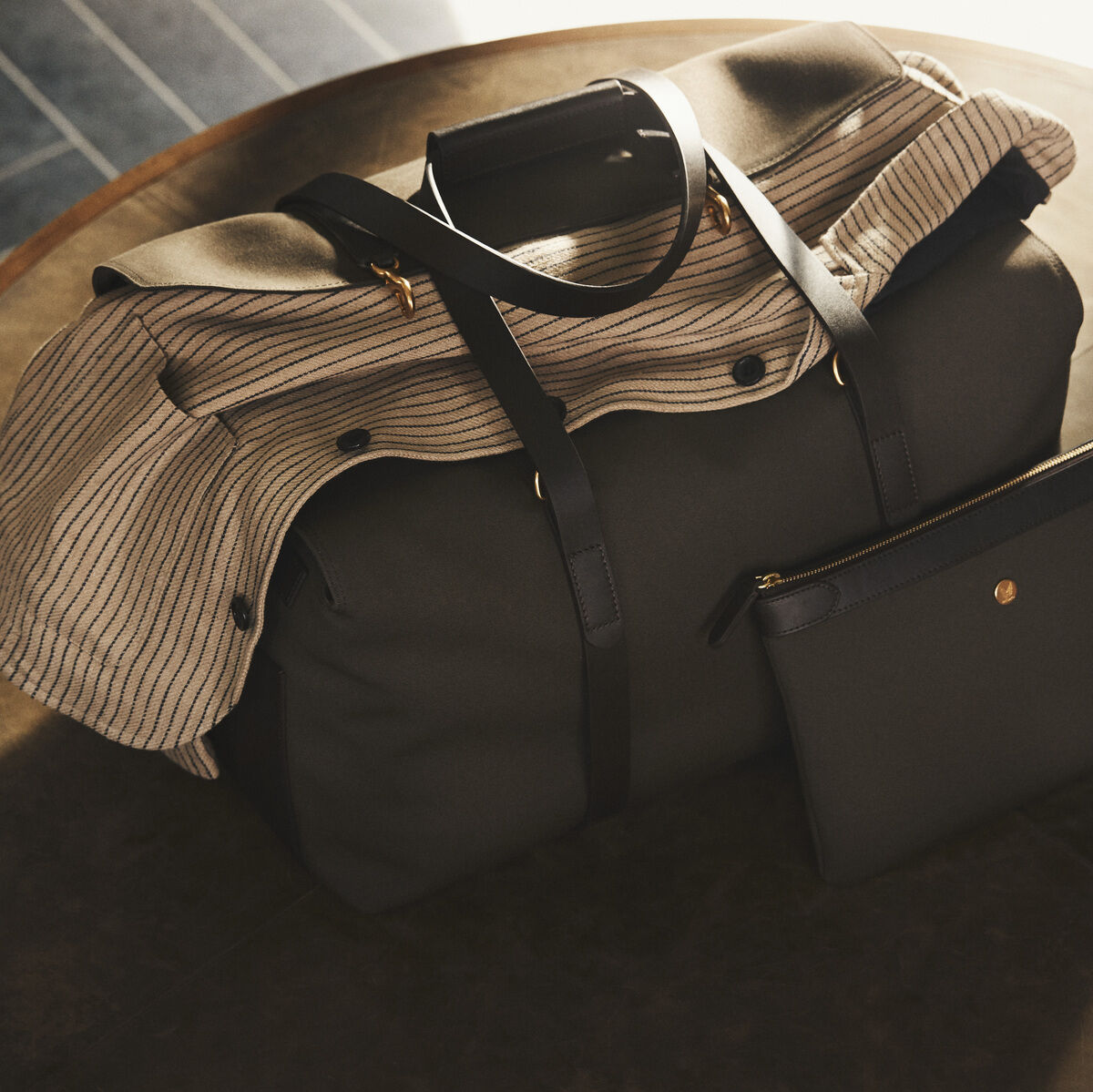Reisetasche »SUPPLY« auf einem Tisch drapiert, begleitet von einer kleineren Tasche. Die Tasche ist aus schwarzem Stoff mit braunen und gestreiften Akzenten und hat goldfarbene Details.