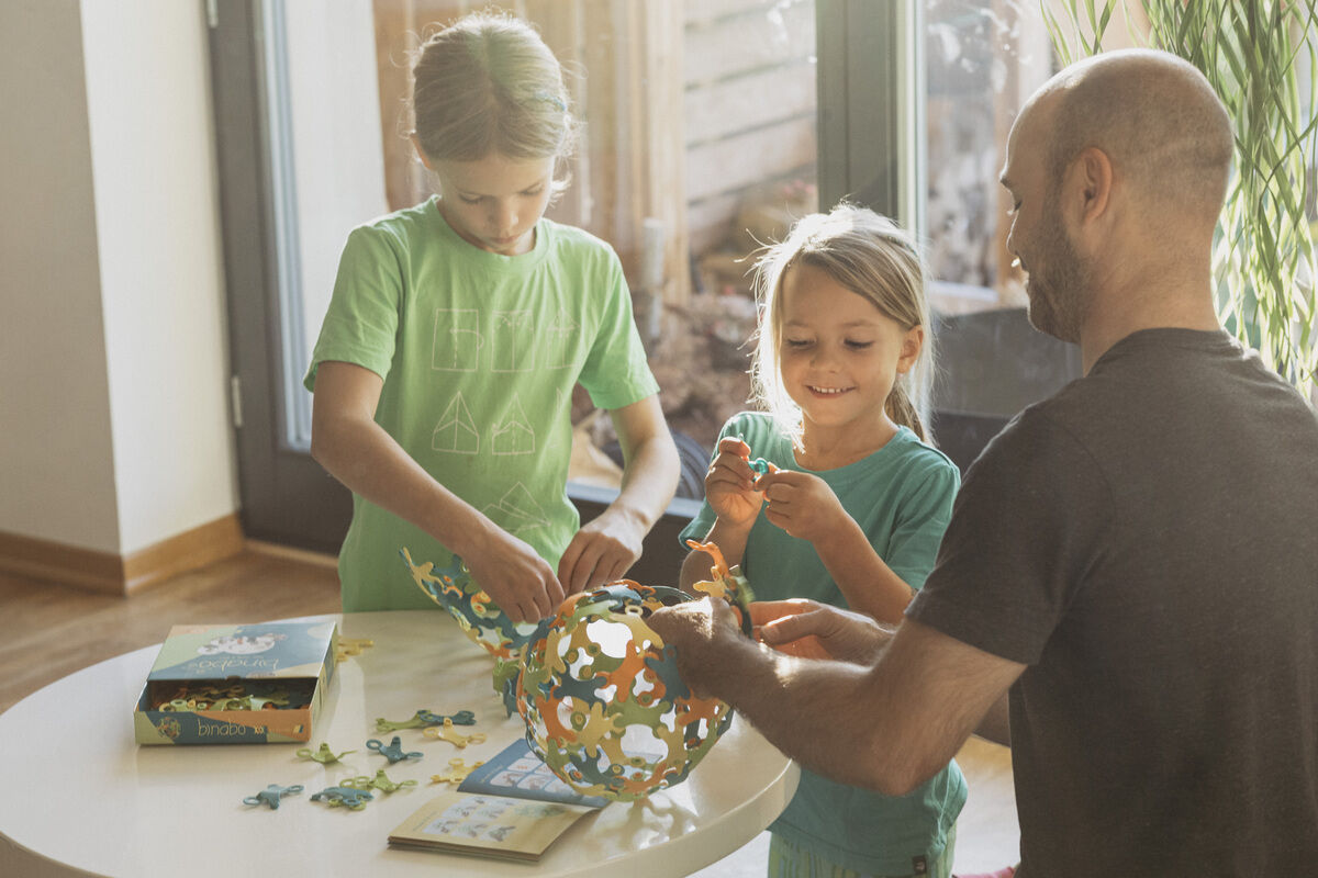 Ein Vater und zwei Kinder spielen gemeinsam mit einem Spiel namens 'Binabo', das aus 60 Teilen besteht. Sie sitzen an einem Tisch und bauen eine konstruktion mit verschiedenfarbigen Teilen.