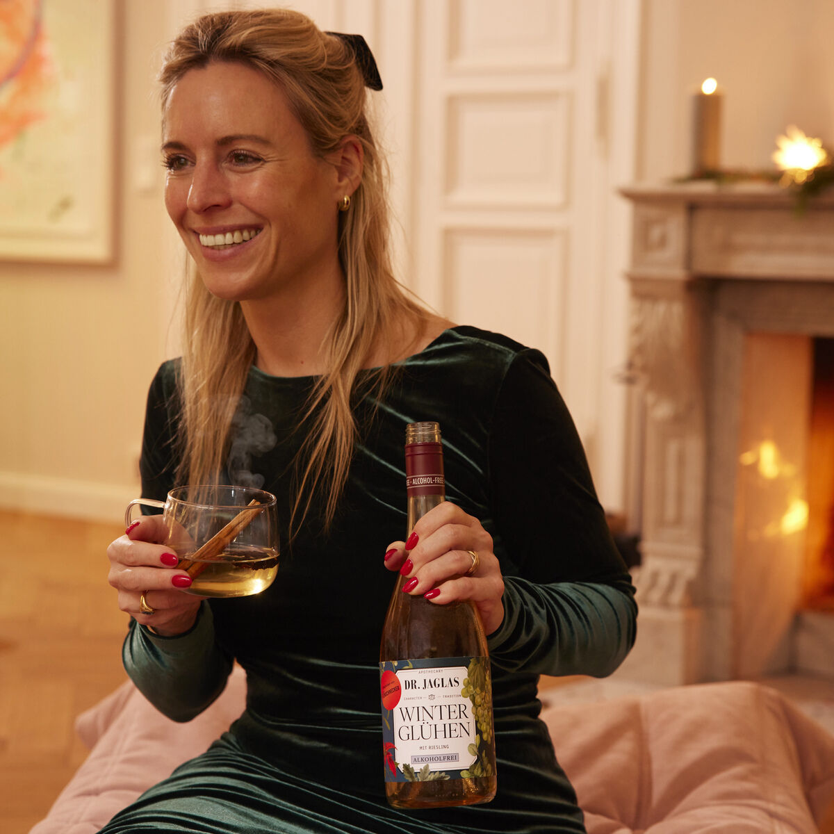 Eine lächelnde Frau in einem grünen Samtkleid sitzt entspannt mit einer Tasse dampfendem Glühwein und einer Flasche "Winterglühen Riesling, 3er Winzerglühwein alkoholfrei" in einem gemütlichen Wohnzimmer vor einem brennenden Kamin, weihnachtliche Dekoration auf dem Kaminsims.