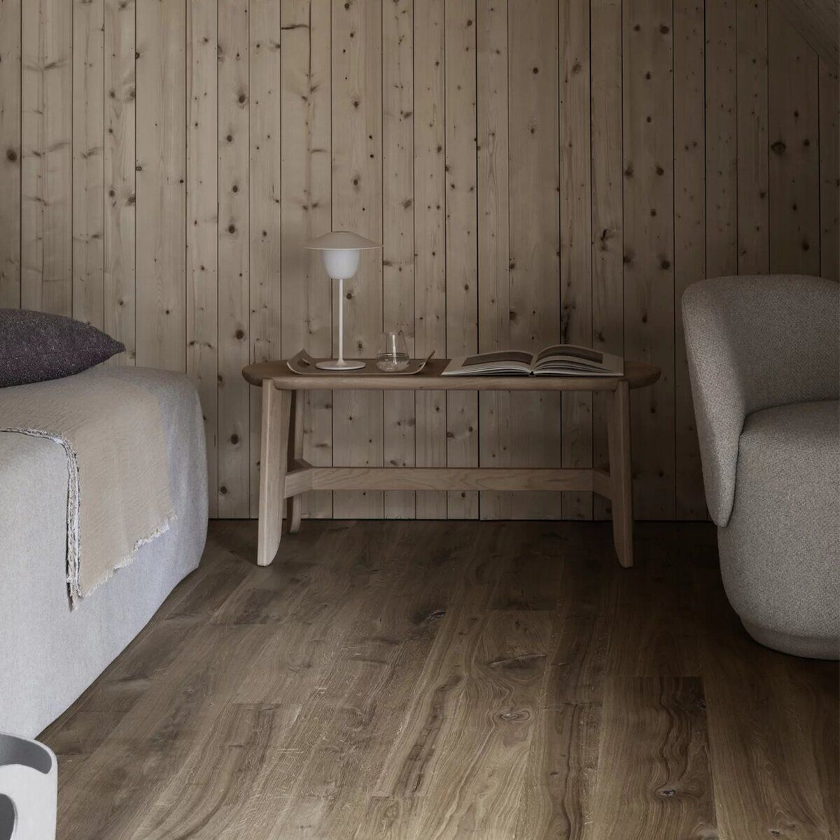 Eine minimalistisch gestaltete Sitzbank aus hellem Holz steht in einem gemütlichen, mit Holz vertäfelten Raum neben einem Bett mit neutraler Bettwäsche und einem modernen Sessel. Auf der Sitzbank befinden sich eine Lampe, zwei Gläser und ein aufgeschlagenes Buch. Die Einrichtung wirkt skandinavisch und lädt zum Entspannen ein.