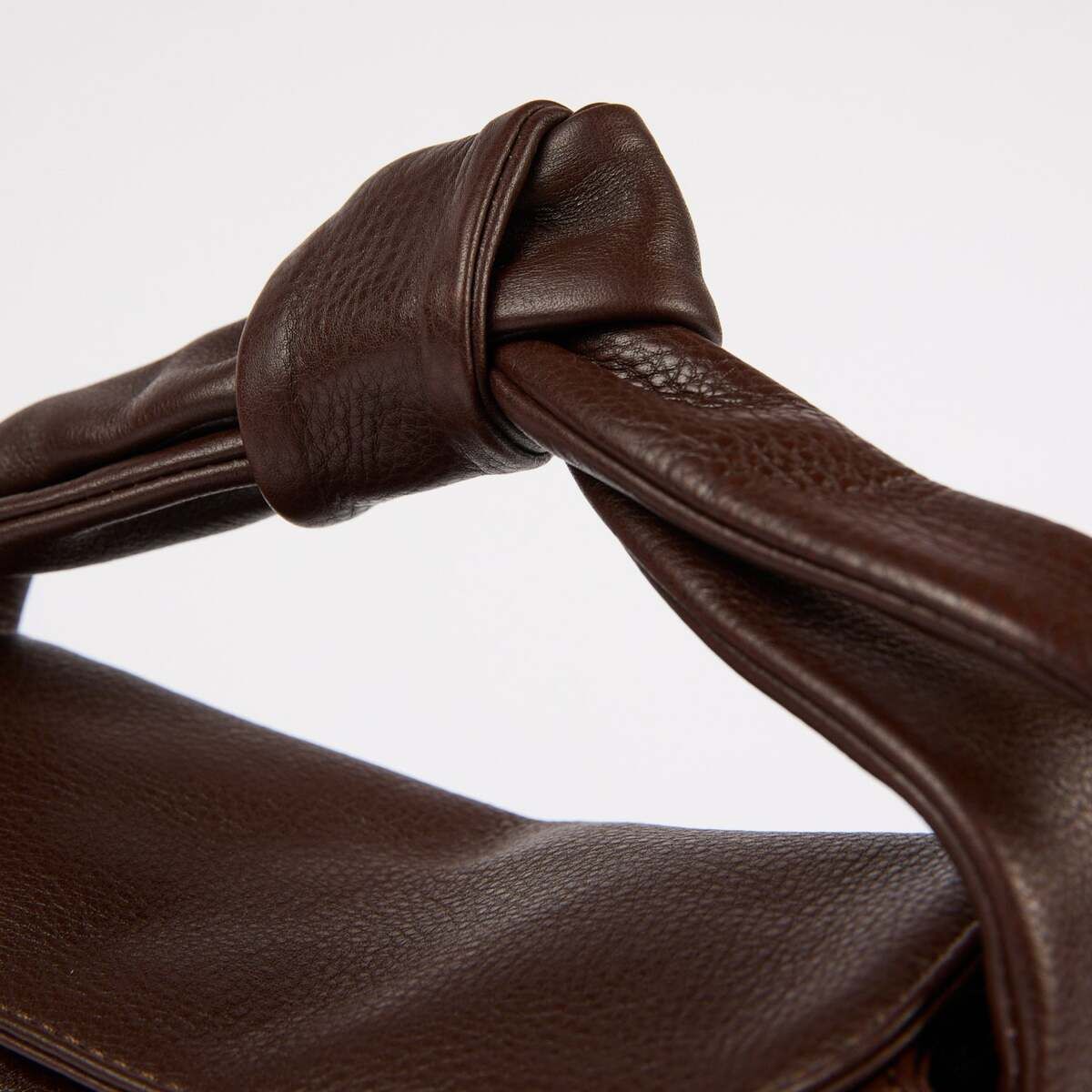 Detailaufnahme einer dunkelbraunen Umhängetasche aus Leder mit Knoten-Detail am Griff. Die Tasche »Coco Croissant« präsentiert sich in elegantem, minimalistischem Design auf neutralem Hintergrund.