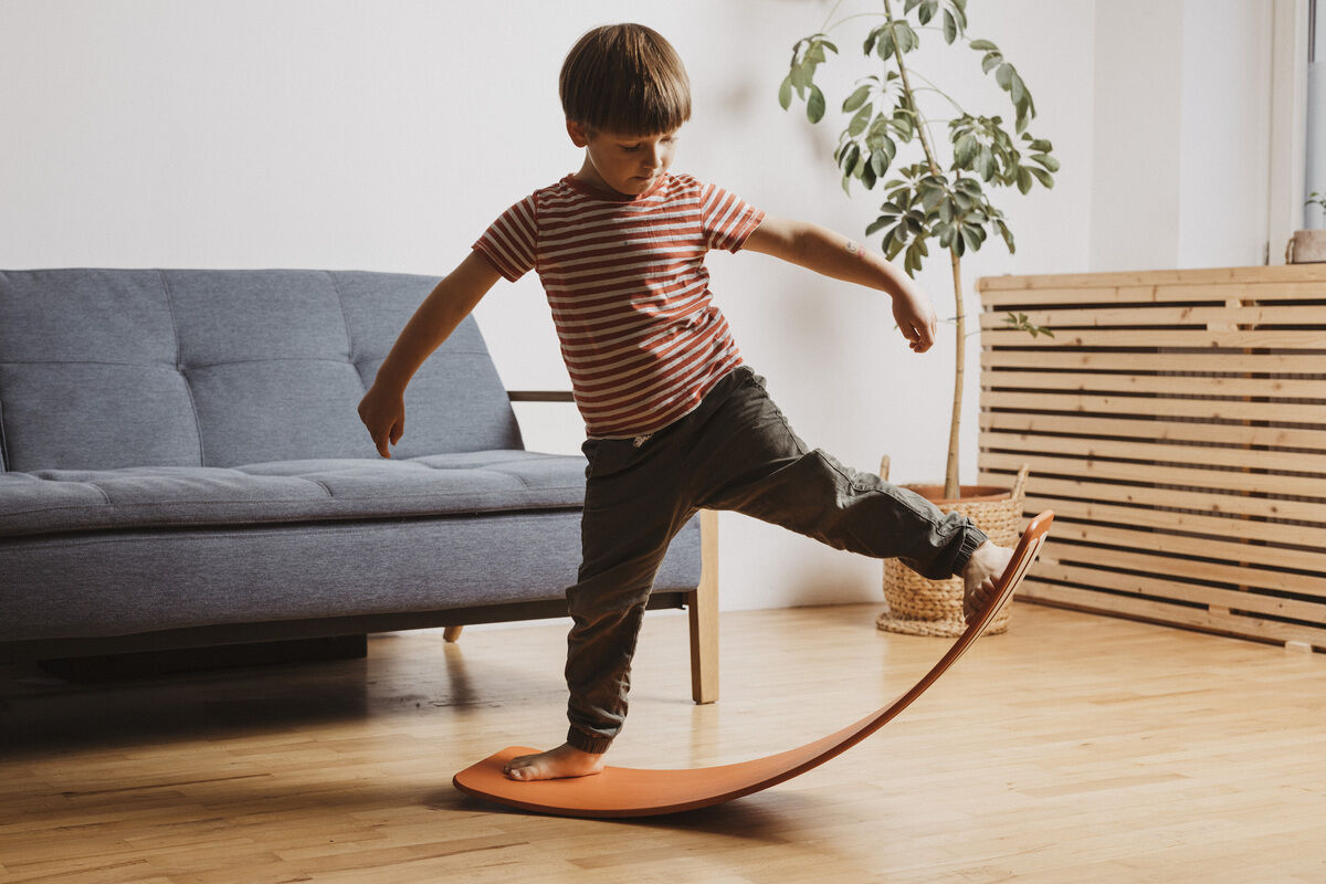 Ein Kind balanciert auf einem roten Balanceboard im Wohnzimmer, das mit einem grauen Sofa, einem Holzboden und einer Pflanze im Hintergrund ausgestattet ist.