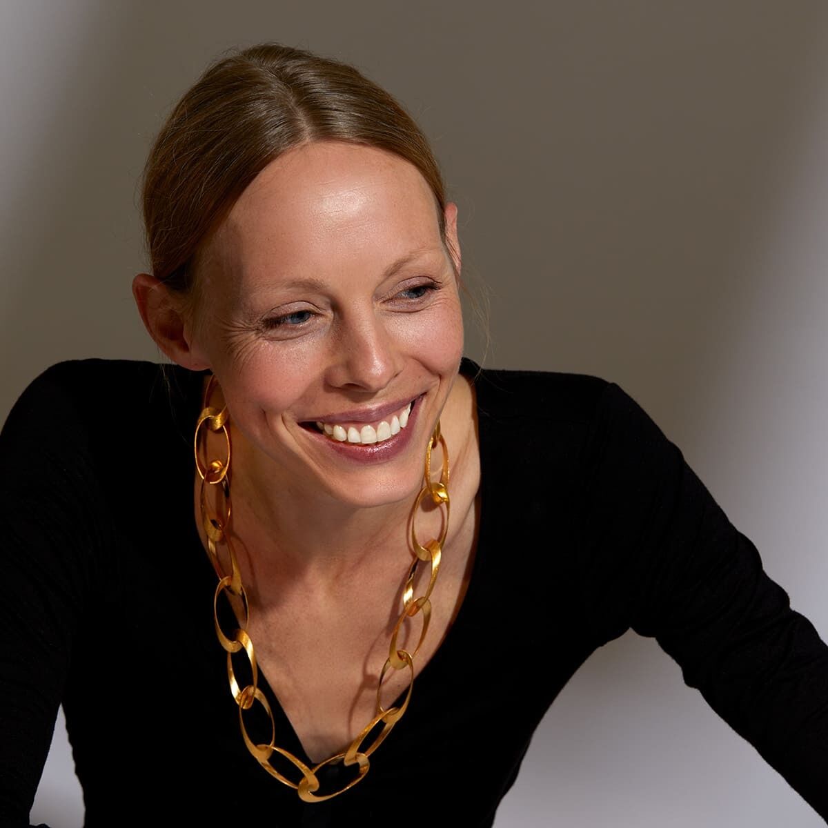 Eine lächelnde Frau trägt ein goldenes Collier mit großen Gliedern und einen schwarzen Oberteil vor einem neutralen Hintergrund.