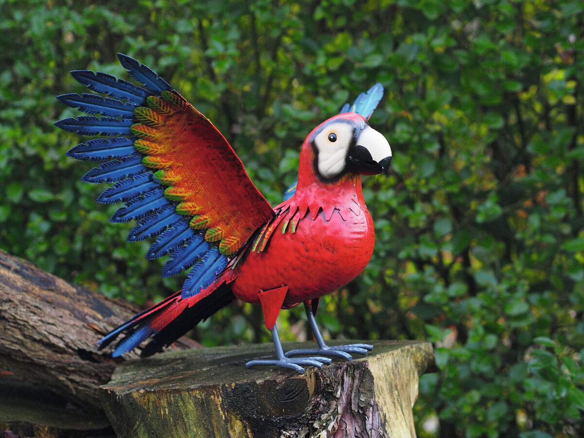 Bunte Gartenfigur eines Papageis aus Metall, die auf einem Baumstumpf im Garten platziert ist. Die Figur ist in leuchtenden Farben wie Rot, Blau, Grün, Weiß und Gelb gehalten, mit detaillierten Merkmalen und ausgebreiteten Flügeln. Der Hintergrund zeigt eine üppige grüne Pflanzenlandschaft.