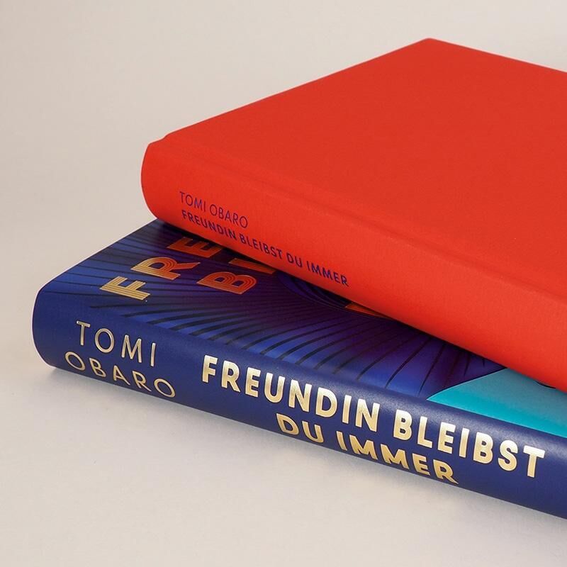 Zwei Bücher liegen übereinander, das obere Buch hat einen roten Einband mit dem Titel und Autorennamen 'Tomi Obaro' geprägt, während das untere Buch einen blauen Einband mit dem Titel 'Freundin bleibst du immer' und dem Autorennamen 'Tomi Obaro' in gelber Schrift hat.