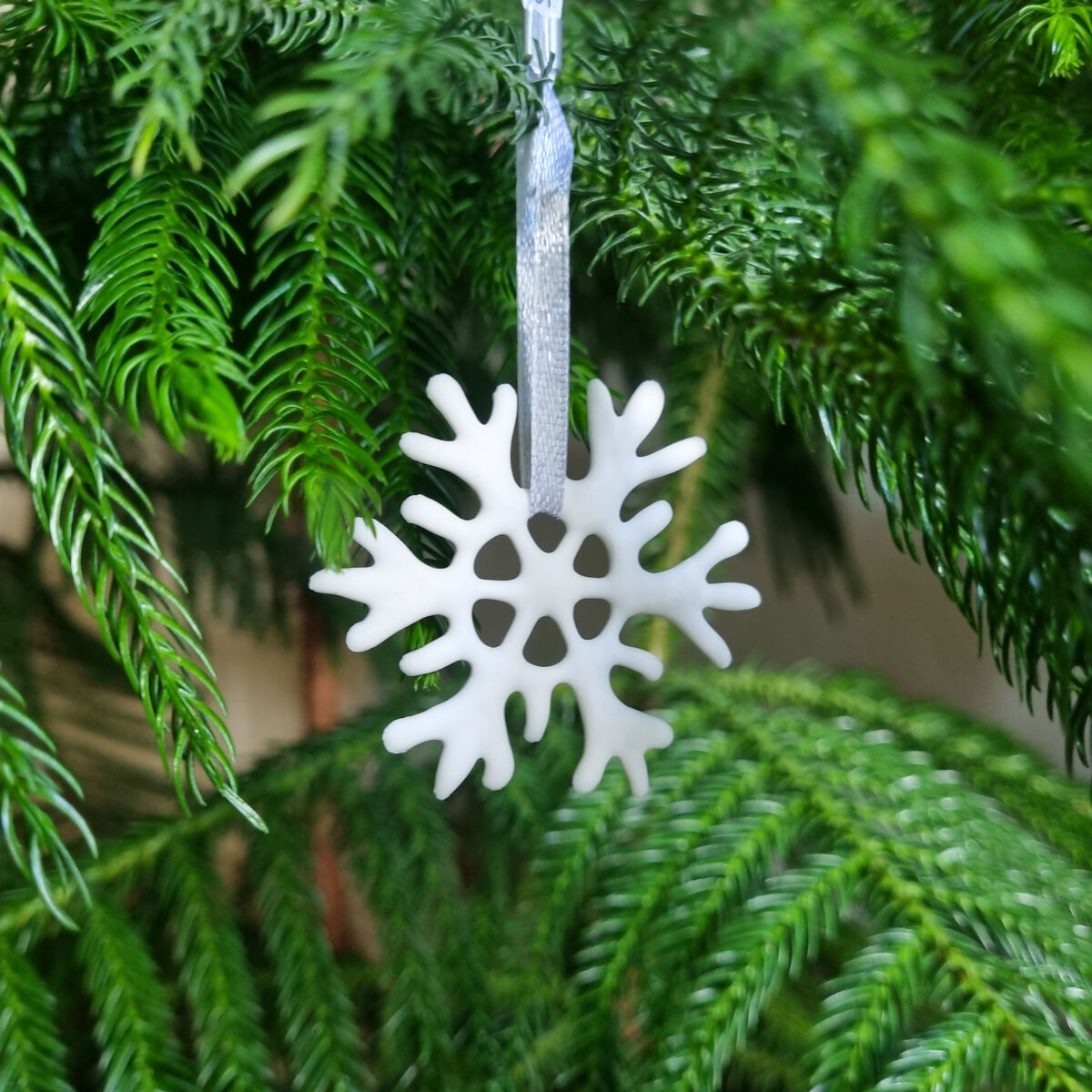 Keramik-Schneeflocke aus dem 8er Set kleine Schneeflocken von Silke Decker hängt an einem silbernen Band an einem grünen Nadelzweig. Die Schneeflocke ist fein gearbeitet, matt weiß und hebt sich kontrastreich von dem satten Grün der Nadeln ab.