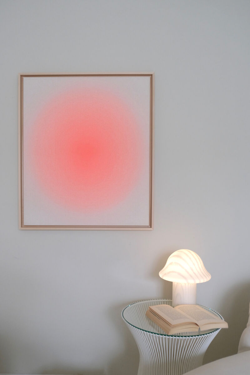 Ein quadratisches Kunstwerk mit einem Farbverlauf in Apricot- und Rosatönen in einem schlichten Rahmen an einer weißen Wand hängend. Darunter steht ein kleiner runder Beistelltisch mit einer Pilzlampen und einem aufgeschlagenen Buch darauf.