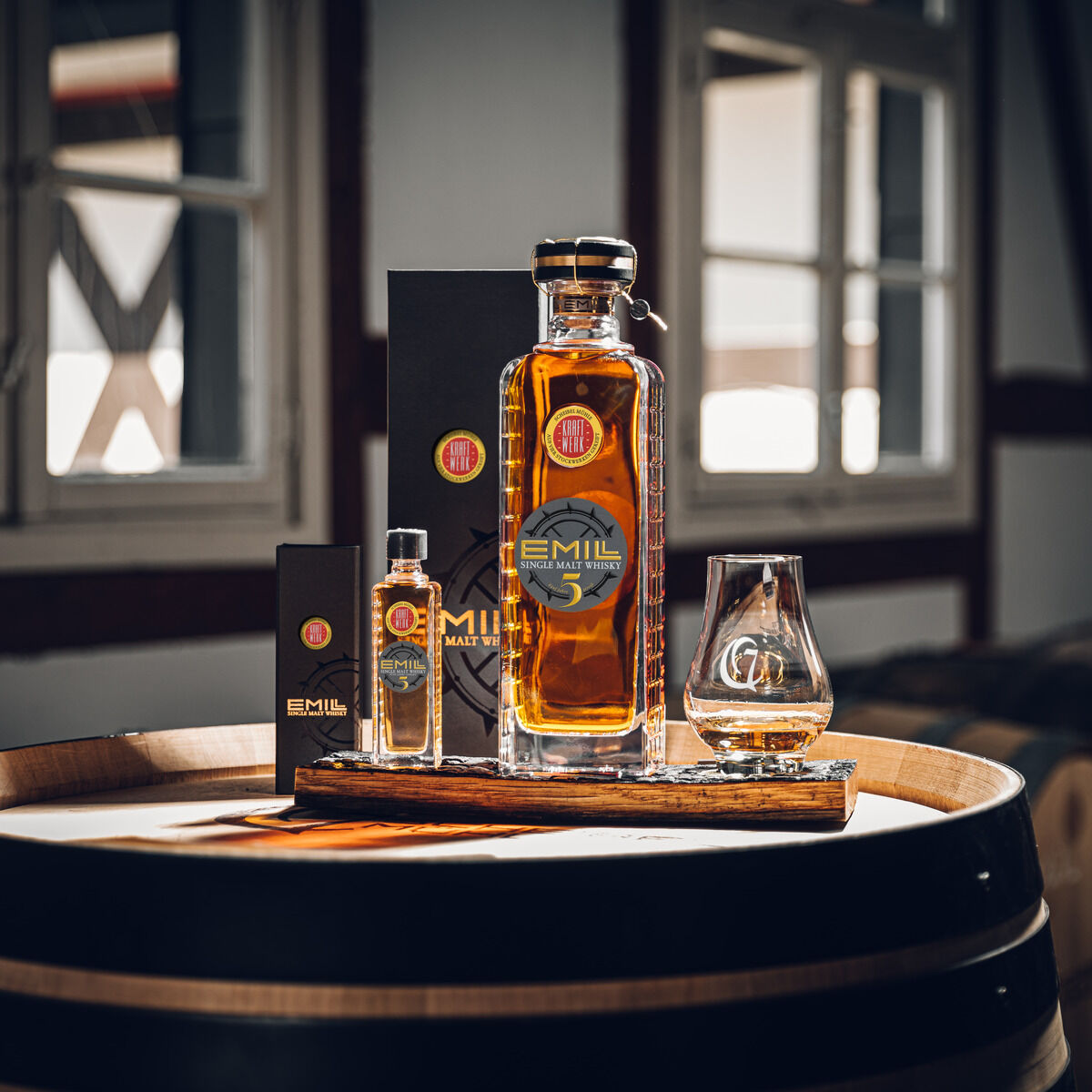 Auf einem Holzfass stehen stilvoll drapiert eine große und eine kleine Flasche Single Malt Whisky "EMILL Kraftwerk" 5 Jahre, dazu ein Whiskyglas und hochwertige schwarze Geschenkverpackungen. Im Hintergrund klassische Fasslagerung und Fenster mit Tageslicht.