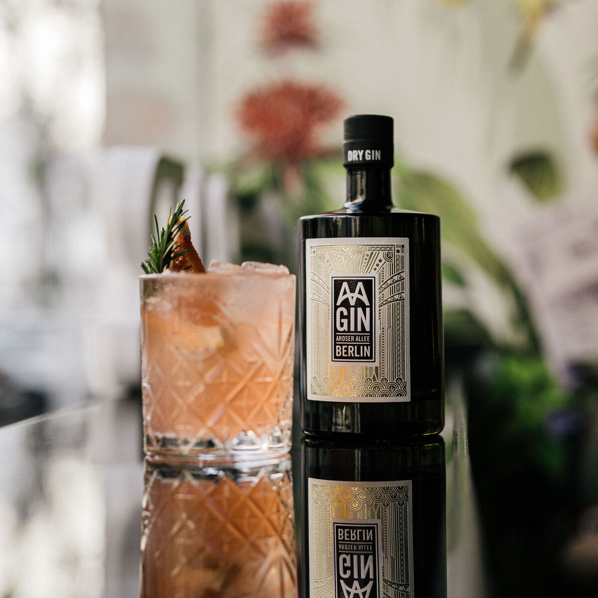 Eine Flasche »AAGIN« Dry Gin im edlen Design von Weindimensional steht neben einem mit Eis gefüllten Kristallglas. Der Drink ist rosa, verziert mit Kräutern und Frucht, im Hintergrund sind florale Muster und verschwommene Pflanzen zu sehen. Alles wirkt hochwertig inszeniert.
