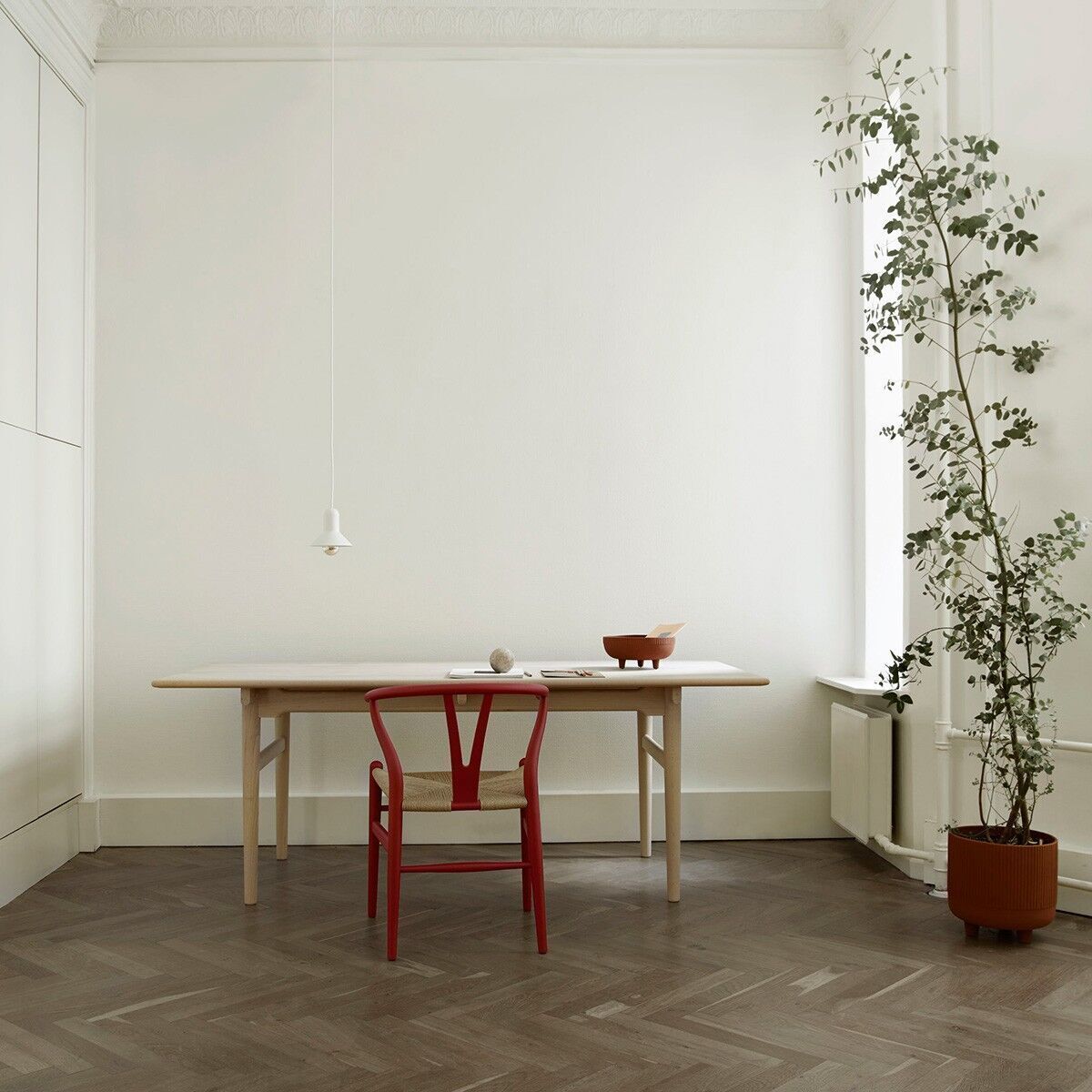 Esszimmer mit einem modernen Tisch und einem einzelnen roten Stuhl aus der Sonderedition »Stuhl CH24 Soft« von Carl Hansen. Im Hintergrund steht eine Pflanze neben einem Fenster, und ein minimalistisches Hängeleuchtelicht hängt über dem Tisch.