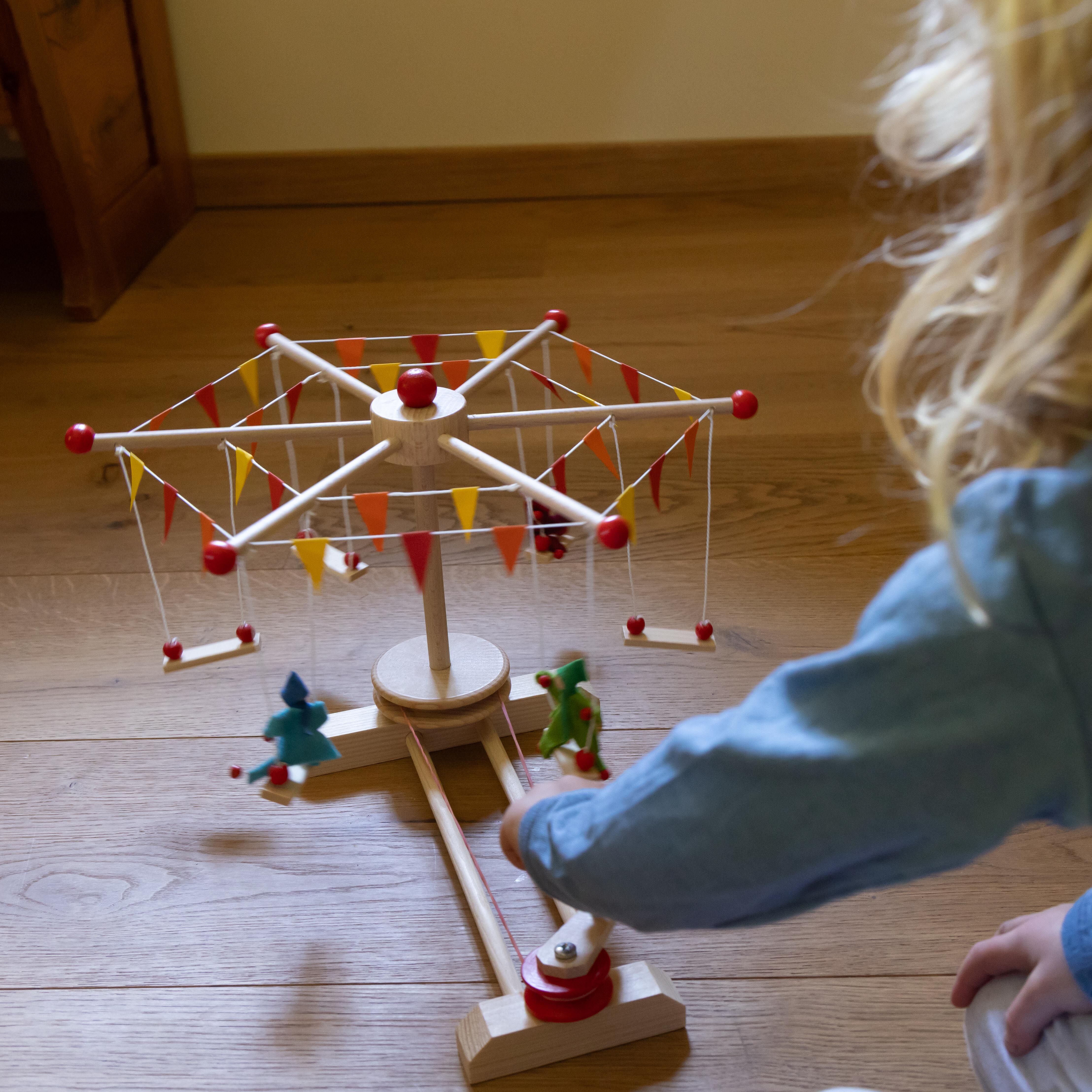 Ein zusammengebautes Kettenkarussell als Holzspielzeug steht auf einem Holzboden. Zwei bunte Holzfiguren sitzen in den Schaukeln des Karussells. Ein Kind berührt das Spielzeug, der Hintergrund zeigt einen Teil eines Kinderzimmers mit Holzmöbeln. Das Bild betont die verspielte, handgemachte Atmosphäre und vermittelt traditionellen Bastelspaß.