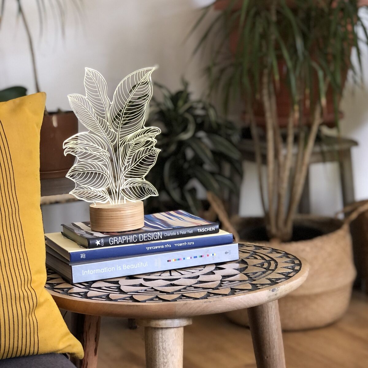 Eine 3D-Lampe in Form von Heliconia-Blättern steht auf einem runden Holztisch vor mehreren Pflanzen, die die Szene umgeben. Die Lampe besteht aus weißen, durchscheinenden Materialien, die bei Beleuchtung ein weiches, warmes Licht abgeben. Der Tisch ist mit einigen Büchern und dekorativen Elementen bestückt. Ein gelbes Kissen und ein Holzboden sind ebenfalls sichtbar.