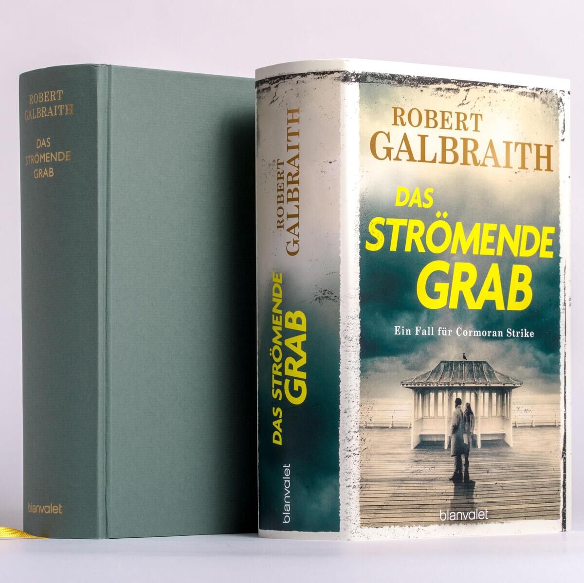 Ein gebundenes Buch mit dem Titel 'Das strömende Grab' von Robert Galbraith ist zu sehen. Der Einband zeigt ein Motiv eines Küstenhauses im Nebel in Gelb- und Grautönen, das auf eine spannungsgeladene Handlung hinweist. Links daneben ist die Rückenansicht des Buches in grünem Leineneinband mit gelbem Lesebändchen abgebildet.