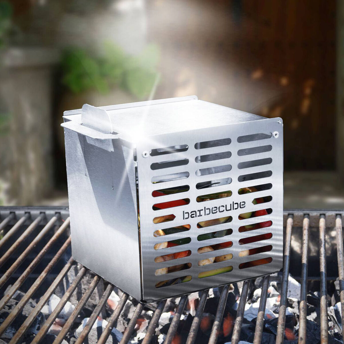 Ein silberner Grillwürfel namens 'The Cube' ist auf einem Grillrost zu sehen. Im Hintergrund befinden sich verschwommene Gartenpflanzen und eine dunkle Holztür. Lichtstrahlen fallen auf den Grill, was den Raucheffekt betont.