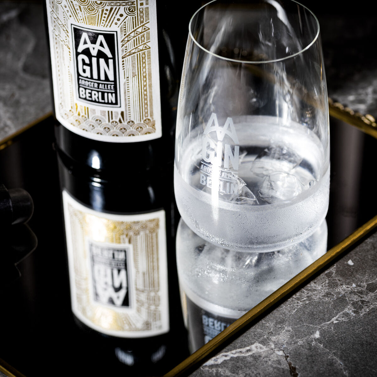 Eine Flasche »AAGIN« Dry Gin und ein Glas im Set präsentiert auf einer reflektierenden Oberfläche mit eleganter, goldener Umrandung, ideal für luxuriöse Anlässe.