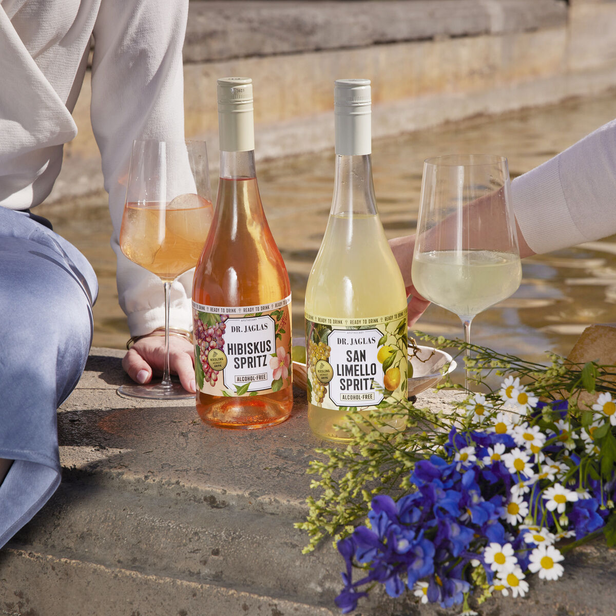 Zwei Flaschen Hibiskus Spritz neben zwei Gläsern voller Getränke, mit einem Blumenstrauß im Vordergrund und zwei Personen, die am Ufer sitzen. Die Szene wirkt sommerlich und entspannt.