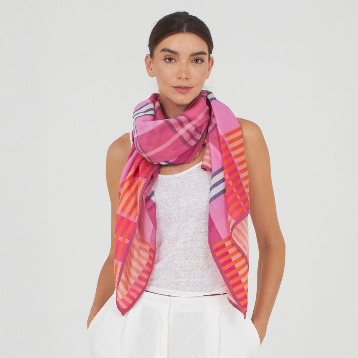 Frau trägt den Baumwoll-Seidenschal »Morgan« von Shalls Barcelona. Der Schal ist großzügig, vorwiegend in Pink, Orange, Lila und Rosa mit Streifen und Karomuster. Sie trägt ein weißes Top und weiße Hose vor neutralem, hellem Hintergrund.