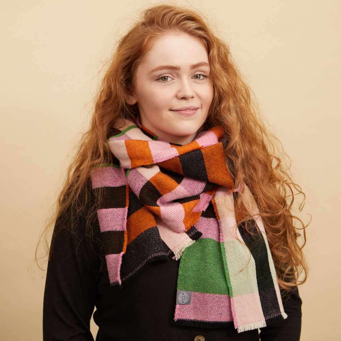 Eine Frau trägt einen bunten Schal oder Wrap von Bauhaus Stölzl. Der Schal weist ein lebendiges Muster in Rosa, Grün, Lila, Orange und Schwarz auf. Sie steht vor einem einfarbigen Hintergrund und trägt eine dunkle Oberbekleidung.