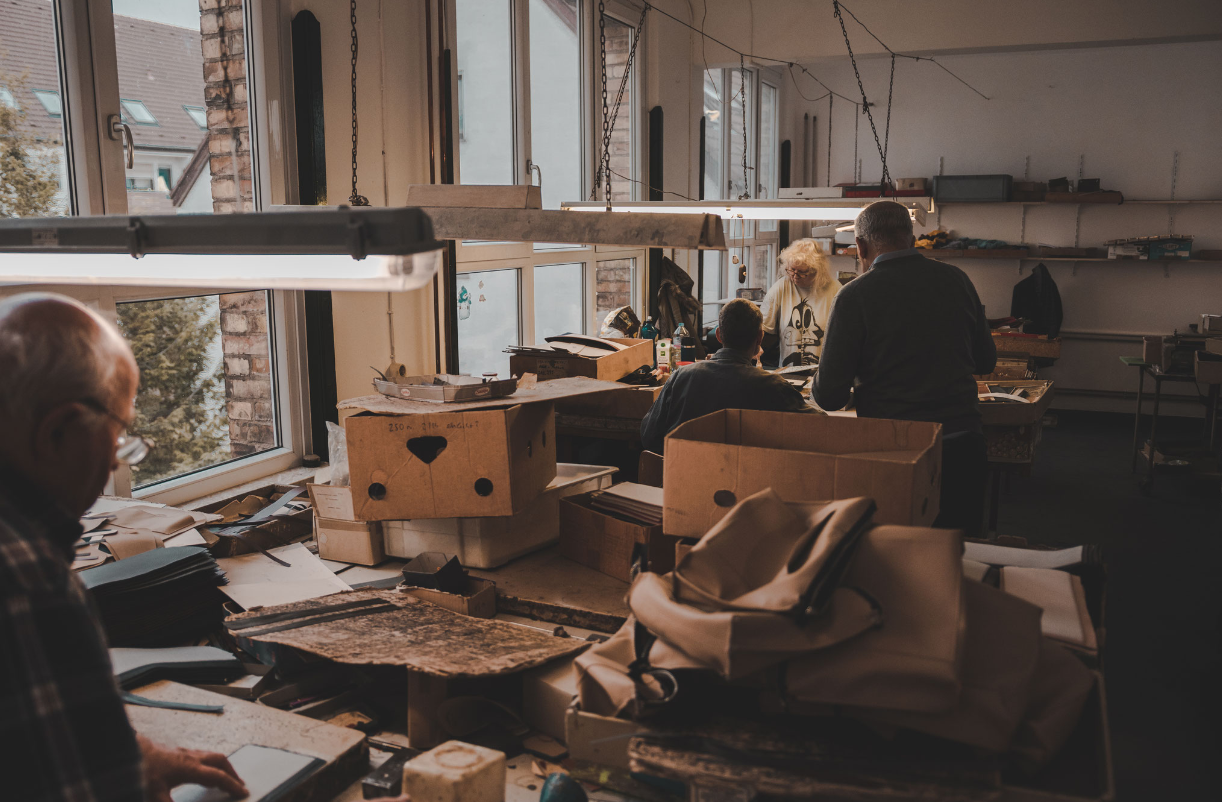 Drei Personen arbeiten in einer Werkstatt mit Holzbearbeitungsmaschinen und Werkzeugen, umgeben von Holzstücken und Kartons.