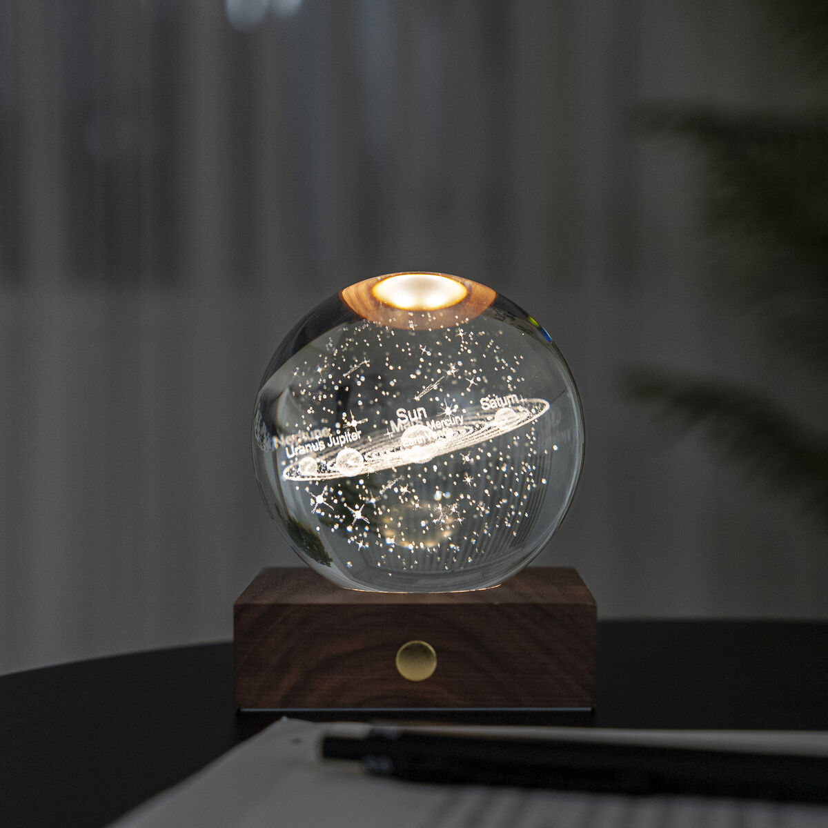 Die Lampe »Amber Light« von Gingko Design steht auf einem edlen Sockel aus Walnussholz. Auf dem Sockel ruht eine kugelförmige Kristallglas-Leuchte mit eingraviertem Sonnensystem, sanft leuchtend. Die Szene wirkt modern und gemütlich, im Hintergrund unscharfe Pflanzen und Vorhänge.