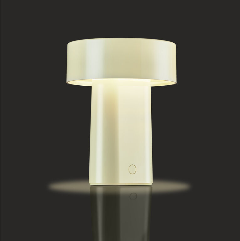 Eine cremefarbene, tragbare Designlampe mit zylindrischem Sockel und breitem, flachem Lampenschirm steht auf einer reflektierenden Oberfläche vor schwarzem Hintergrund. Die Lampe leuchtet in einem warmweißen Licht.