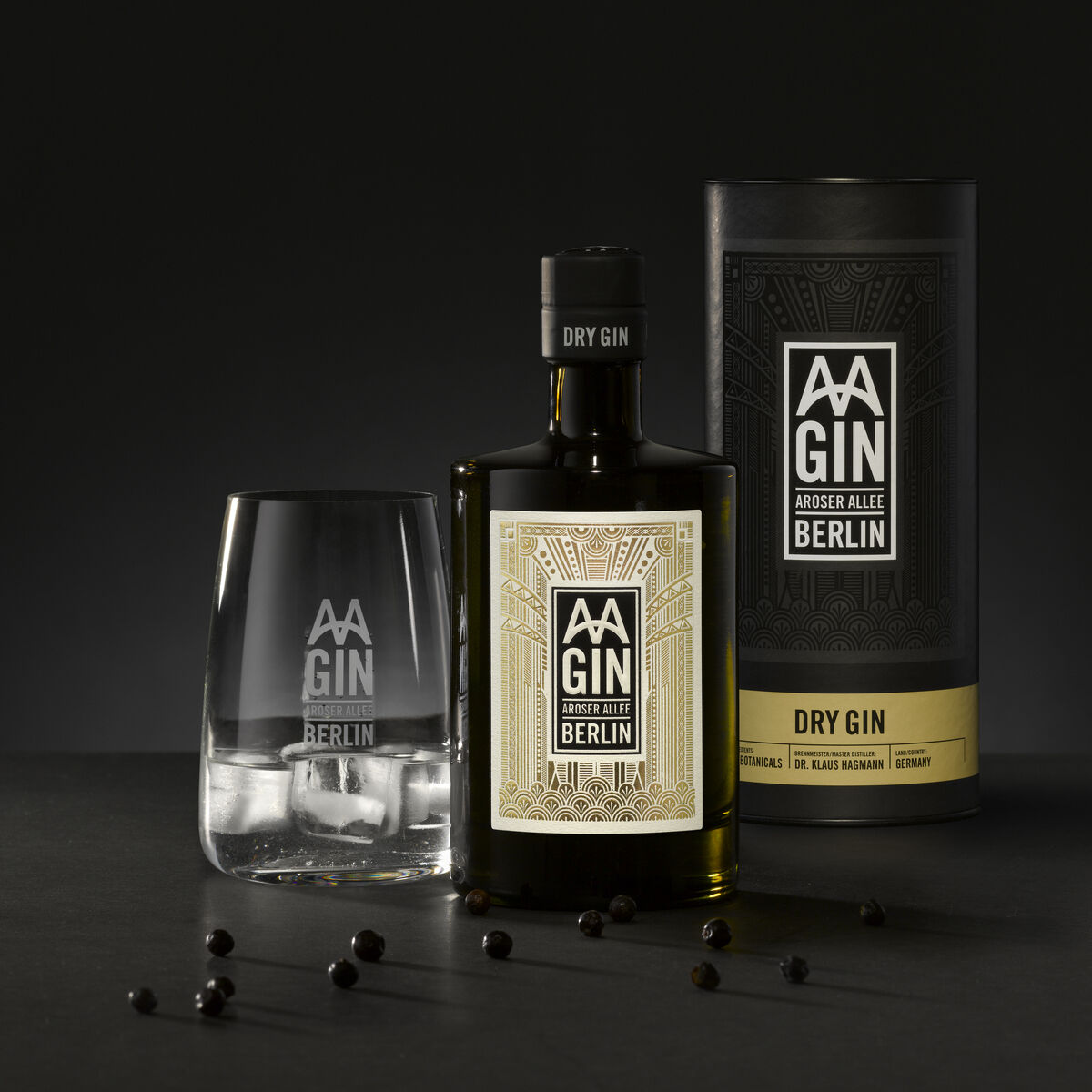 Ein Set aus »AAGIN« Dry Gin und einem dazugehörigen Glas. Der Gin wird in einer eleganten Flasche und einer Tube-Verpackung präsentiert. Einige Wacholderbeeren sind um das Set verstreut. Das Bild hat einen dunklen Hintergrund.