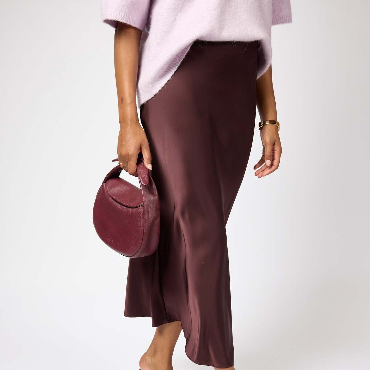 Frau hält eine kleine, runde Handtasche namens »Coco Croissant« in Bordeaux-Farbe. Sie trägt einen fließenden, braunen Rock und elegante, dunkelrote Absatzschuhe. Die Szene vermittelt einen modernen, stilvollen und minimalistischen Look für festliche oder alltägliche Anlässe.