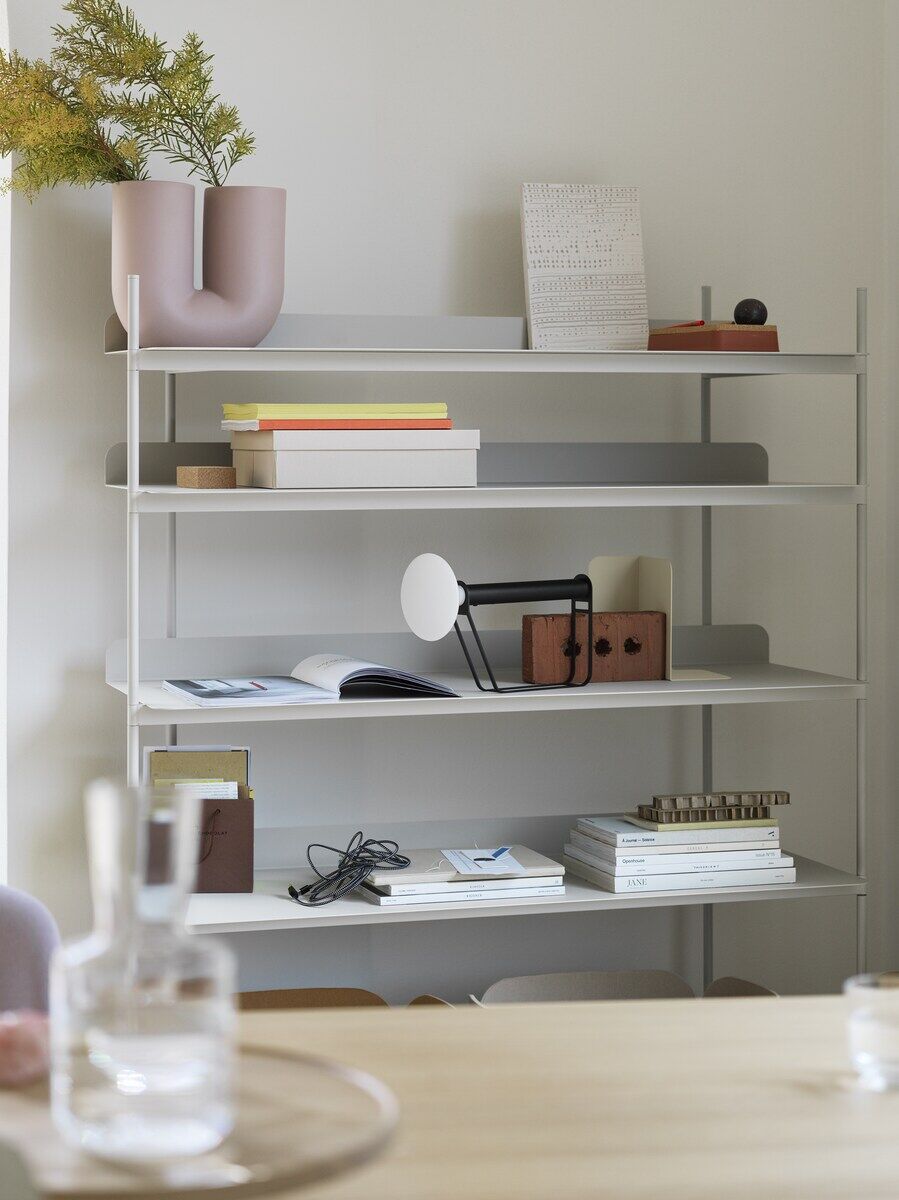 Ein Bücherregal mit mehreren Artikeln, darunter eine tragbare schwarze und weiße Lampe namens »Piton«, Bücher, Pflanzen und Deko-Objekte. Die Lampe steht im mittleren Regal und hat ein modernes, minimalistisches Design.
