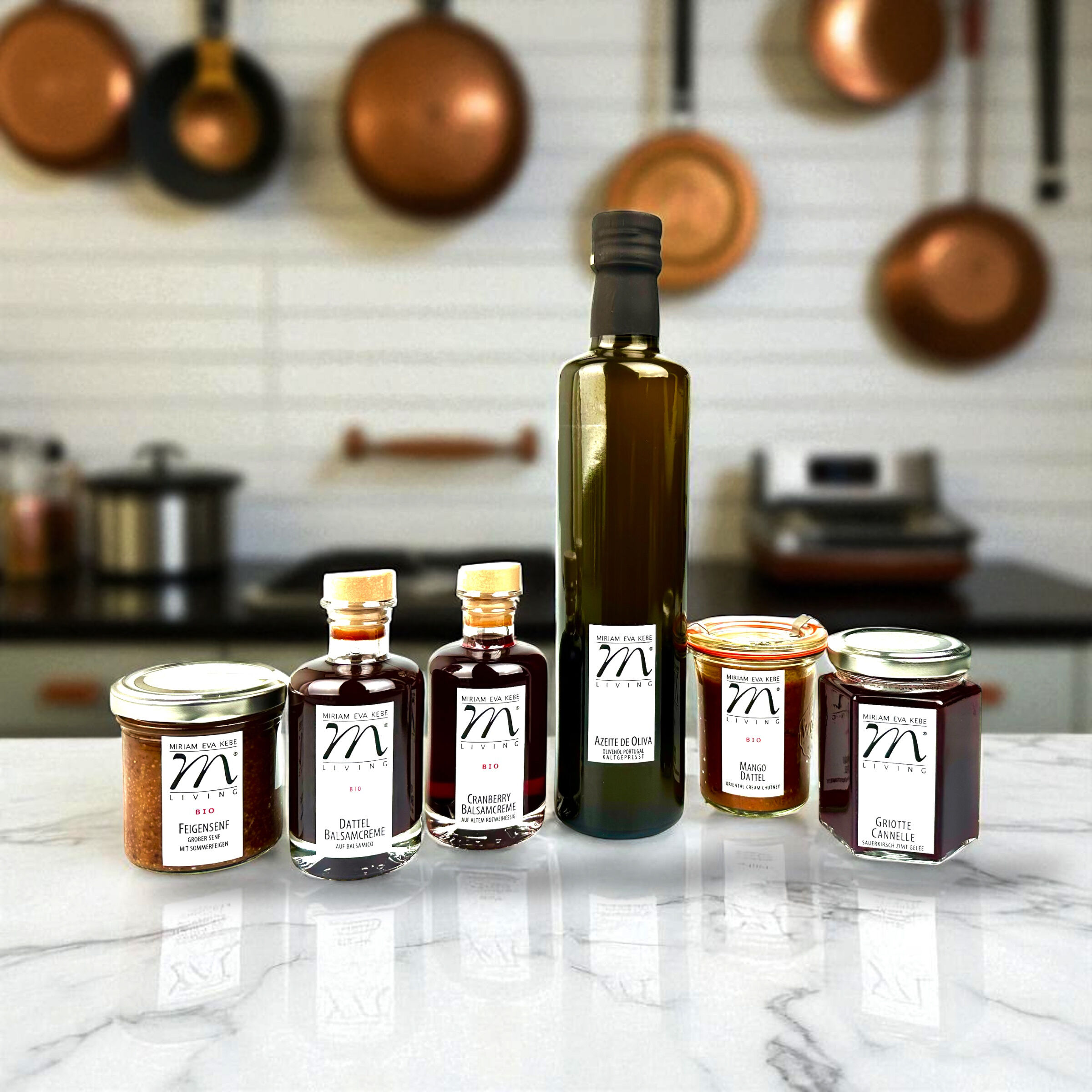 Auf einer hellen Arbeitsplatte steht ein Set aus sechs verschiedenen Gourmet-Produkten in Gläsern und Flaschen, darunter Olivenöl, Essig, Marmelade, Chutney und Gewürzcreme. Die Produkte sind hochwertig etikettiert. Im Hintergrund sieht man eine moderne Küche mit Kupfertöpfen an der Wand.