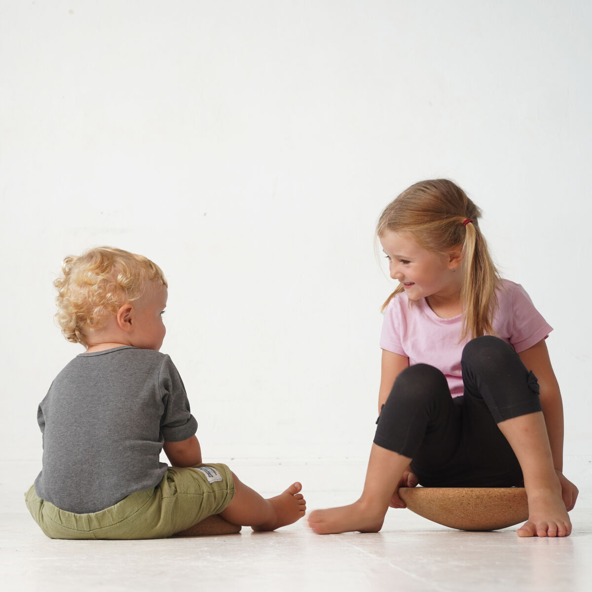 Zwei Kinder spielen mit einem Holzspielzeug in einem hellen Raum. Ein kleines Kind sitzt auf dem Boden, während das andere Kind auf einem Balancierspielzeug sitzt und lächelt.