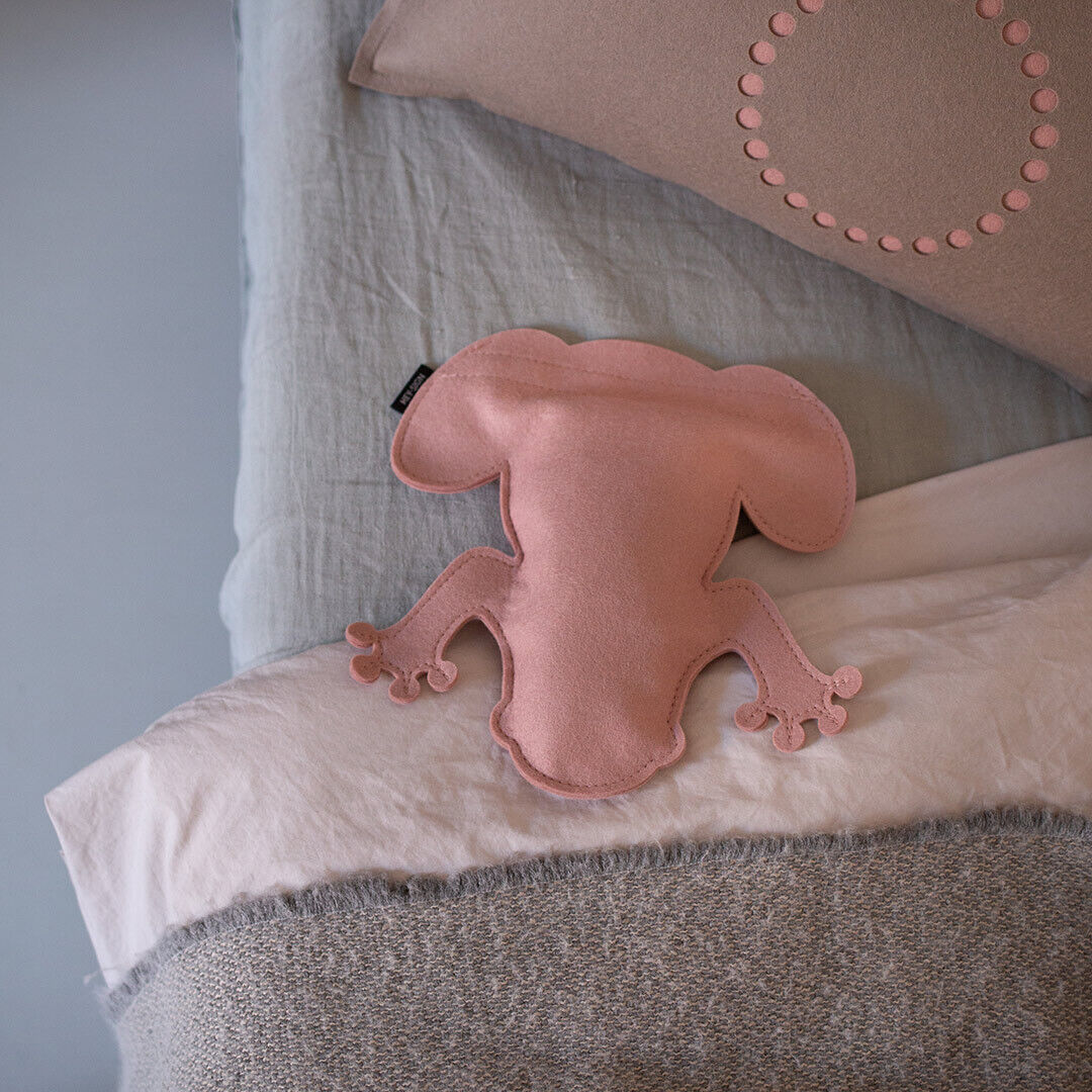Auf dem Bild sitzt ein rosa Kirschkernkissen in Form eines Frosches namens »Frosch Nico« auf mehreren übereinandergelegten Kissen in Beige, Weiß, Grau und Blau.
