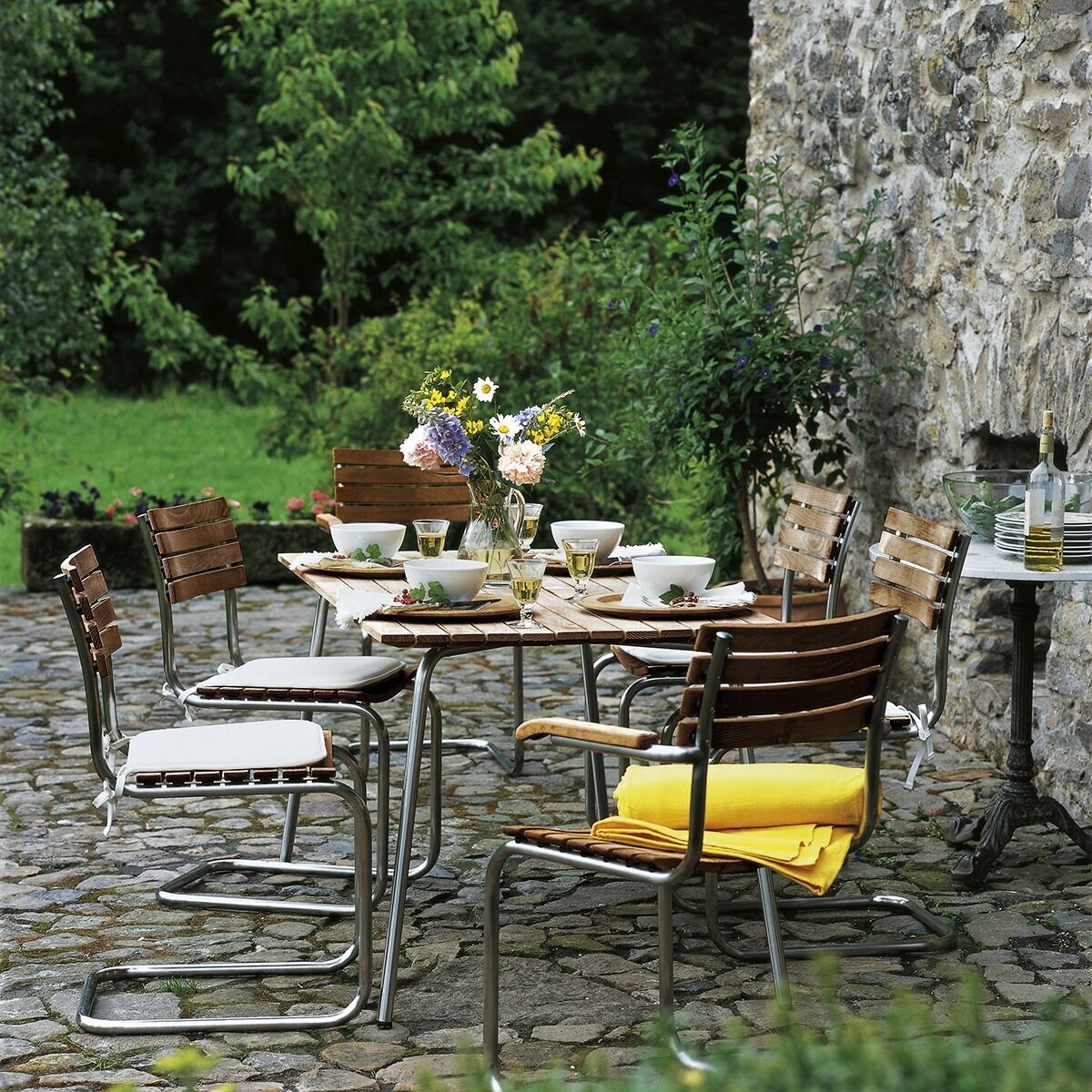 Ein Gartentisch von Thonet, Modell S 1040, umgeben von Holz- und Metallstühlen, auf einer Terrasse mit Kopfsteinpflaster und einer Steinwand. Es ist mit Geschirr und Blumen dekoriert und im Hintergrund ist grüne Vegetation sichtbar.