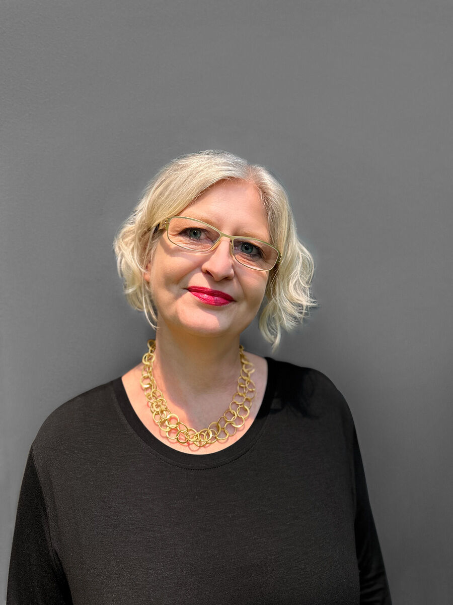 Eine Frau trägt ein »Double«-Collier aus vergoldetem Silber. Sie trägt eine schwarze Oberbekleidung und eine Brille und hat kurzes blondes Haar. Der Hintergrund ist grau.