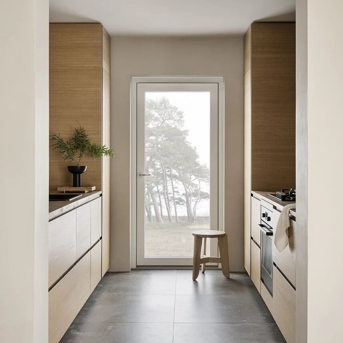 Heller Küchenraum mit modernen, schlichten Holzmöbeln im skandinavischen Stil, zentral steht ein kleiner, naturfarbener Sitzhocker 'ELI' vor einer Glastür mit Blick auf Bäume und Natur, dekoriert mit einer grünen Pflanze in einer schwarzen Vase.