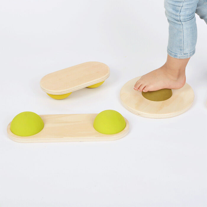 Ein Set von Balanciersteinen aus Holz, bestehend aus verschiedenen Formen mit gelb-grünen Halbkugeln, die Balancier- und Bewegungsübungen für Kinder ermöglichen. Ein Kind im Hintergrund nutzt die Balanciersteine auf einem weißen Hintergrund.