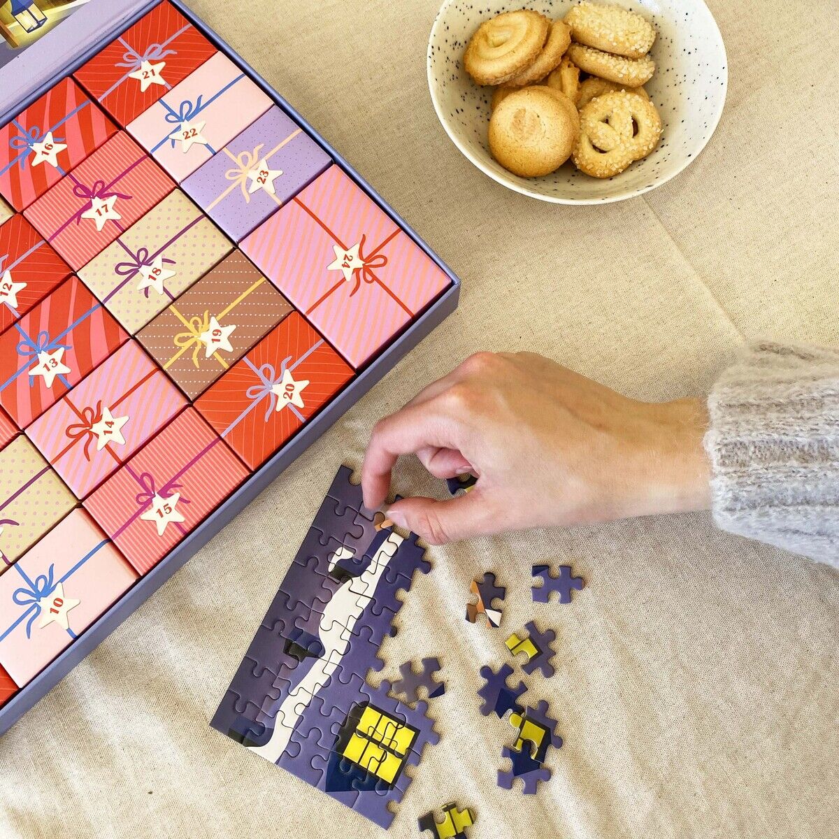 Detailreiche Szene mit dem VISSEVASSE Puzzle-Adventskalender »Skate & Coffee«. Einzelne Adventstürchen in Geschenkoptik, Hand legt Puzzleteile zu violettem Motiv mit gelbem Fenster, Schale mit Buttergebäck, Postkarten mit Weihnachtsbaum auf beigem Tischtuch.