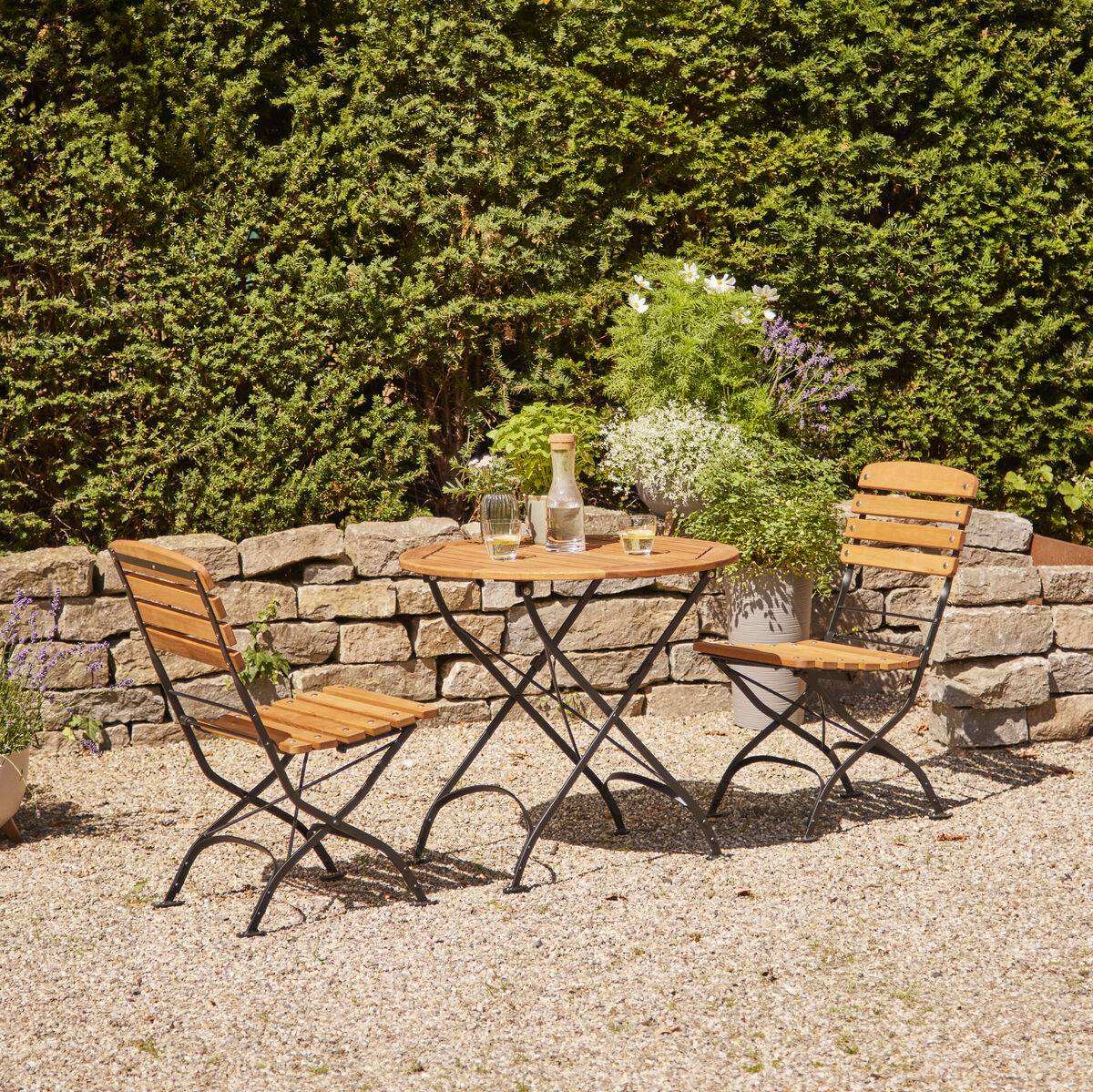 Outdoor-Terrasse mit einem runden Tisch und zwei Klappstühlen aus Holz und Metall. Umgeben von grünen Hecken und dekorativen Pflanzen in Kübeln. Kiesboden und sonniges Wetter.