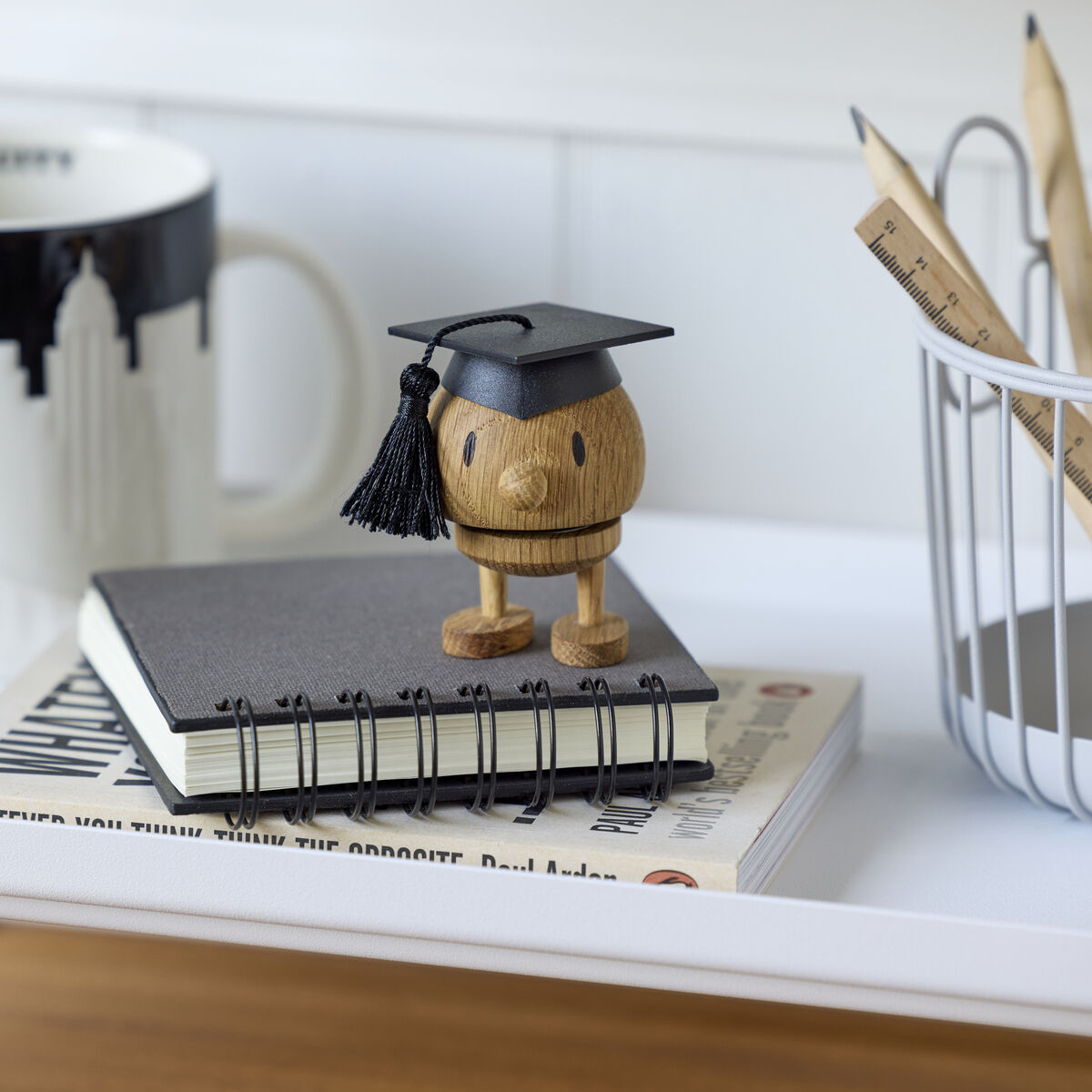 Auf einem weißen Tisch stehen eine Kaffeetasse mit Skyline-Motiv, ein Stiftehalter mit Holzstiften, zwei Bücher und die Holzfigur IQ Bumble Hoptimist mit Doktorhut. Die Szene wirkt aufgeräumt und ist im minimalistischen Design gehalten.