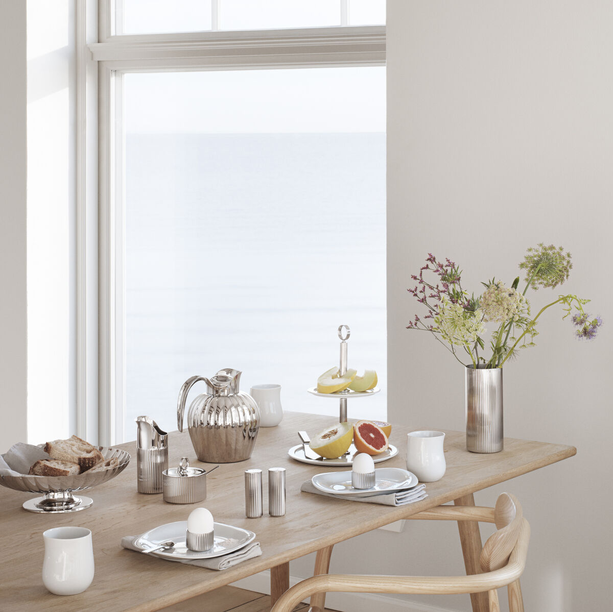 Ein stilvoll eingerichteter Tisch in einem Esszimmer, gedeckt mit Geschirr, darunter ein Sahnekännchen »Bernadotte«. Das Ambiente wirkt hell und minimalistisch, mit einem Fenster im Hintergrund, das auf das Meer hinausblickt. Auf dem Tisch befinden sich weiße Tassen, eine silberne Teekanne, Teller mit Gebäck und ein Blumenstrauß in einer Vase.
