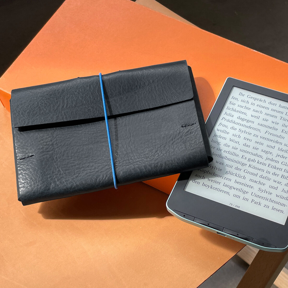 Ein E-Book-Lesegerät in einem schwarzen Etui, das auf einer Oberfläche liegt. Daneben befindet sich eine orangefarbene Ablage oder Verpackung und eine beige Tisch- oder Arbeitsfläche. Ein hellblaues Band sichert das Etui, während das Lesegerät auf einem Holzgestell ruht und seine Display-Texte zeigt.