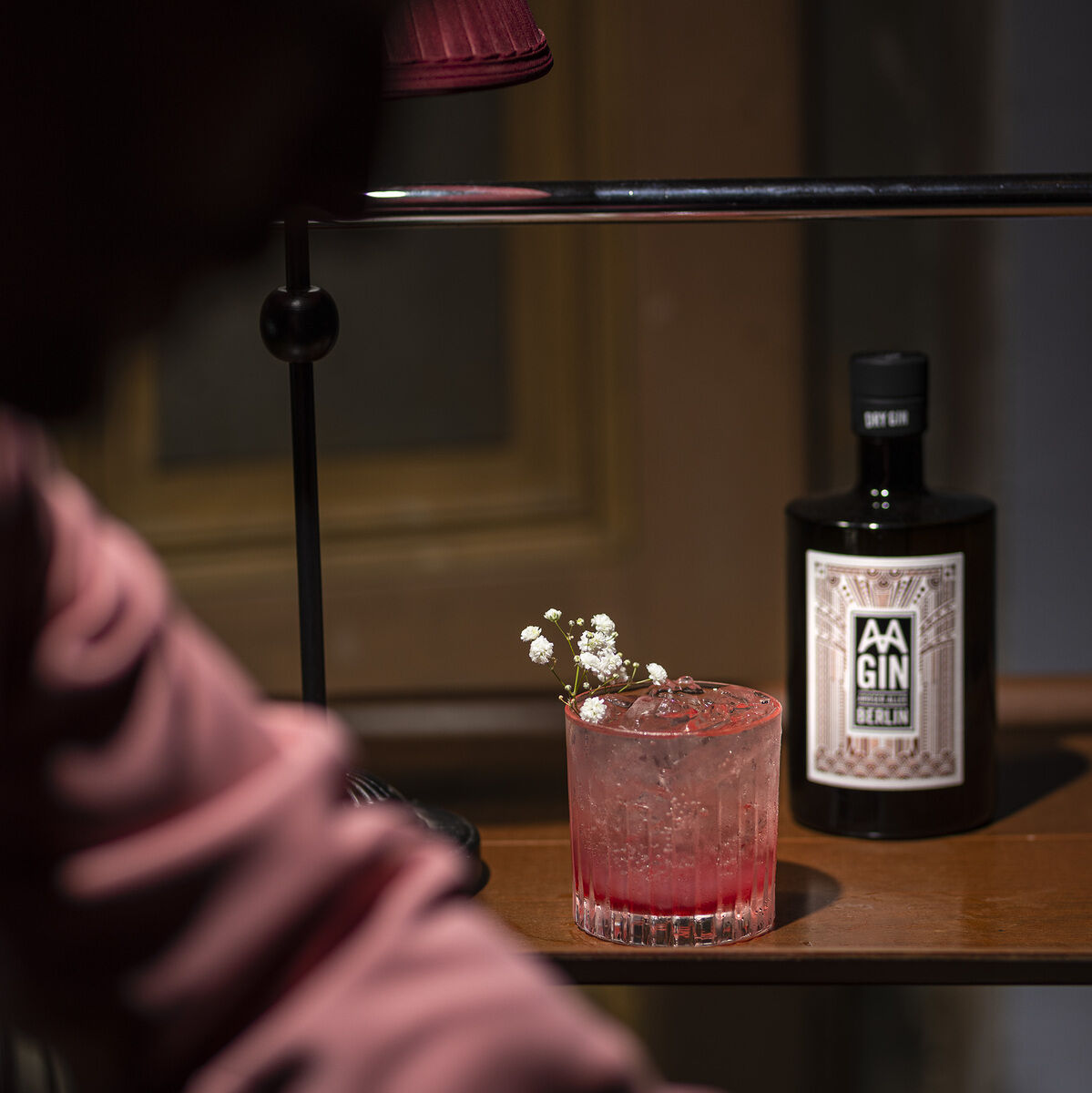 Das Bild zeigt eine Flasche AAGIN Edition 1963 und einen rosafarbenen Cocktail mit Eiswürfeln und weißen Blumen als Dekoration auf einem Holztisch. Im Hintergrund ist eine Person zu sehen.