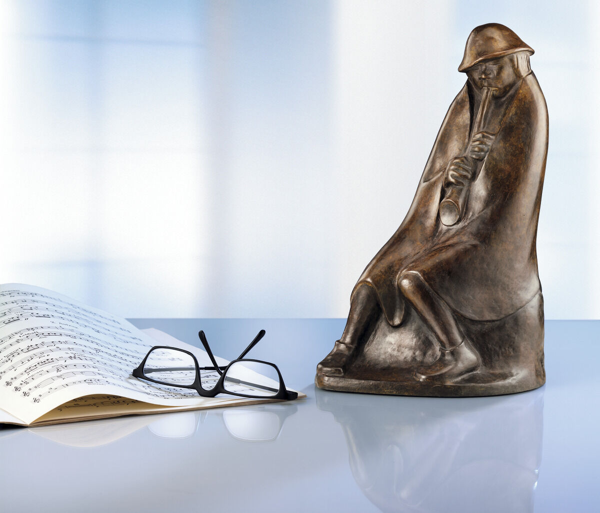 Die Skulptur 'Der Flötenbläser' (1936) von Ernst Barlach zeigt eine sitzende Figur, die eine Flöte spielt. Die Bronze-Skulptur steht auf einer reflektierenden Oberfläche neben einer offenen Partitur und einer Brille. Der Hintergrund ist unscharf und in blauen und weißen Tönen gehalten.