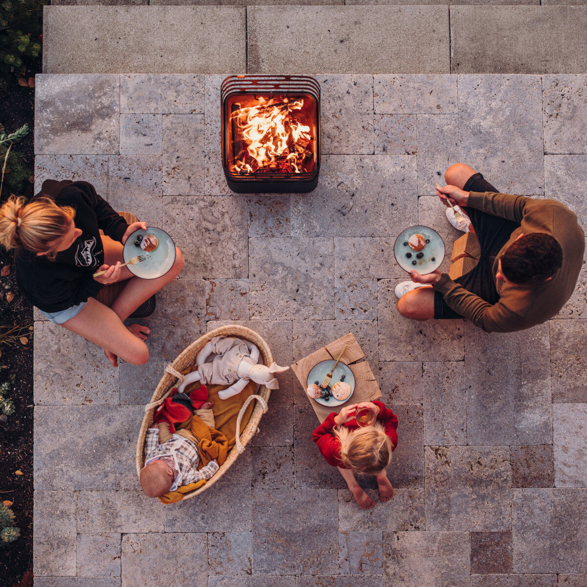 Eine Draufsicht auf eine Terrasse, wo eine Familie um eine Feuerstelle versammelt ist und Mahlzeiten auf Tellern genießt. Eine moderne Plancha ist im Zentrum des Fotos zu sehen.