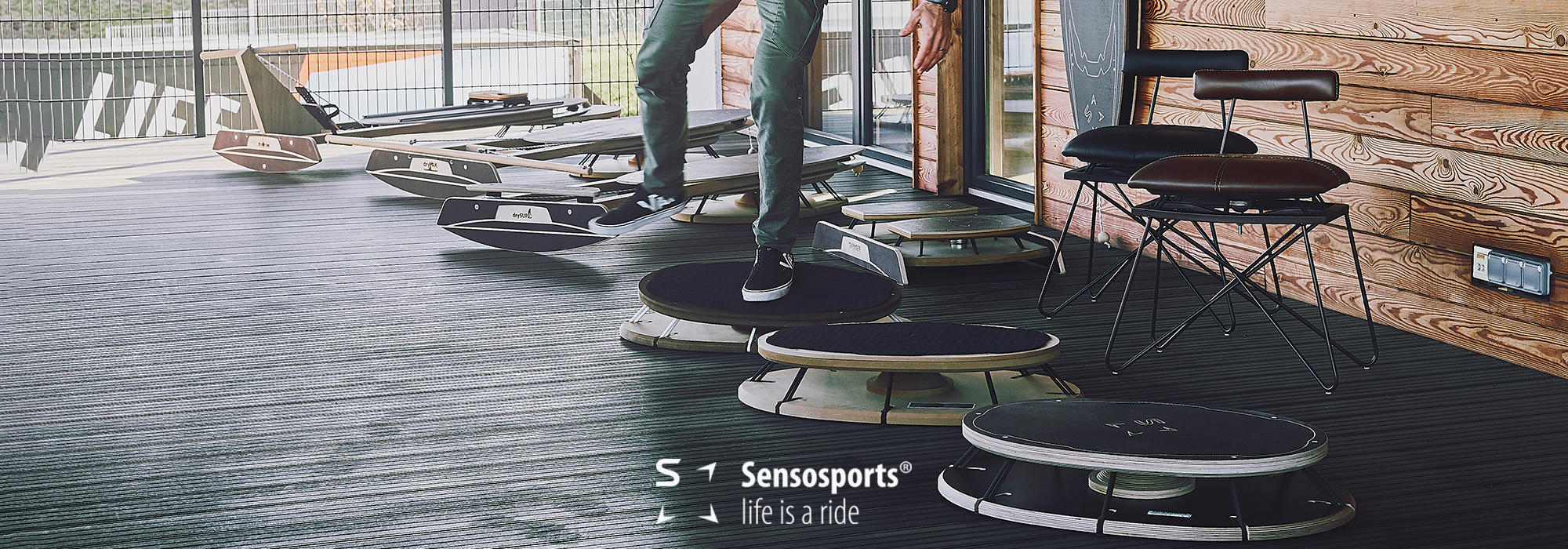 Mehrere schwarze Sensosports® Balance-Boards auf einem Holzboden, eine Person steht auf einem Board, im Hintergrund Holzhauswand und Stühle